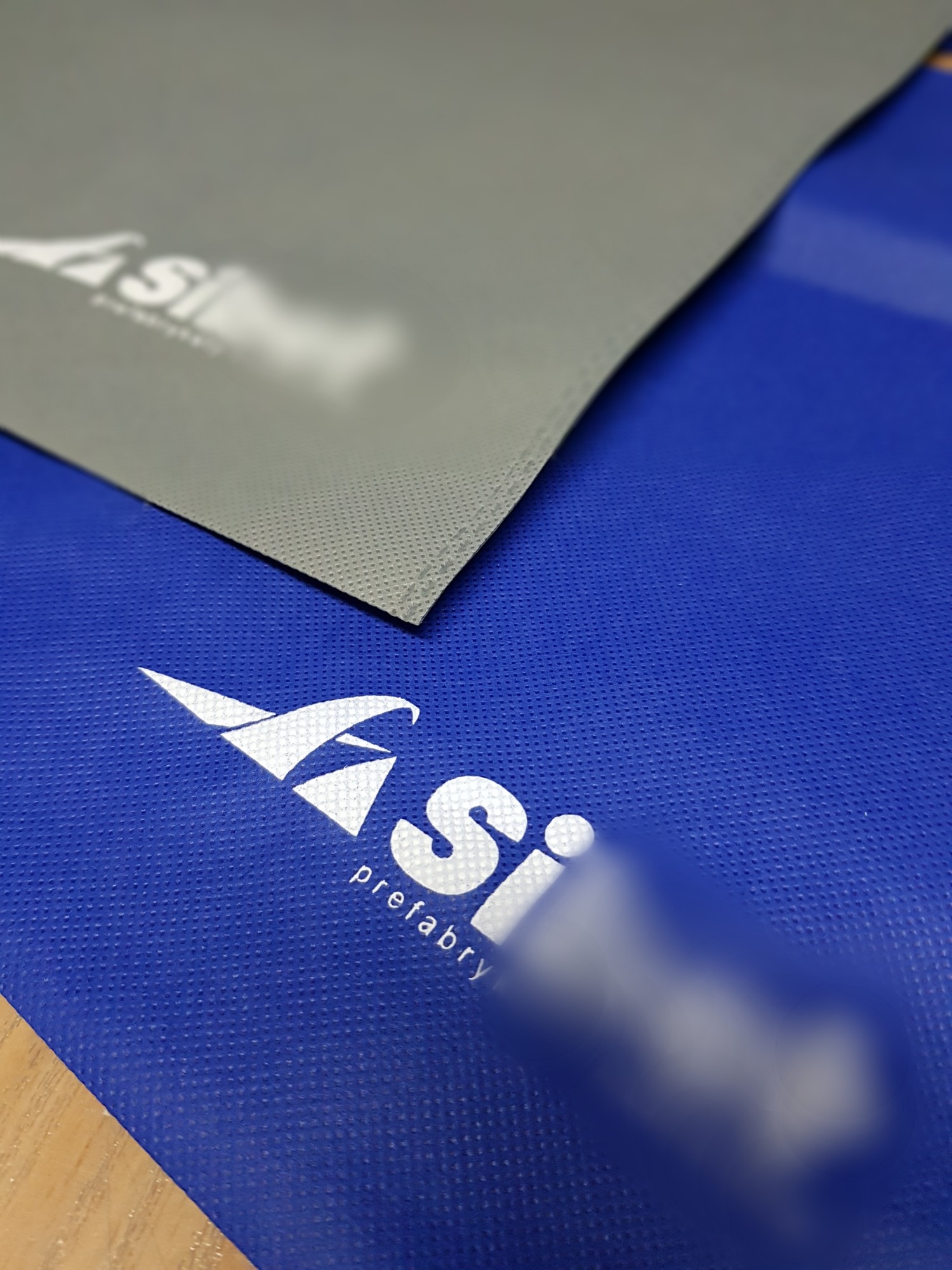 Dwa arkusze materiału z nadrukowanym logo firmy 'Si prefabrykaty', jeden szary z widoczną fakturą, drugi niebieski z delikatnym wzorem kropek.