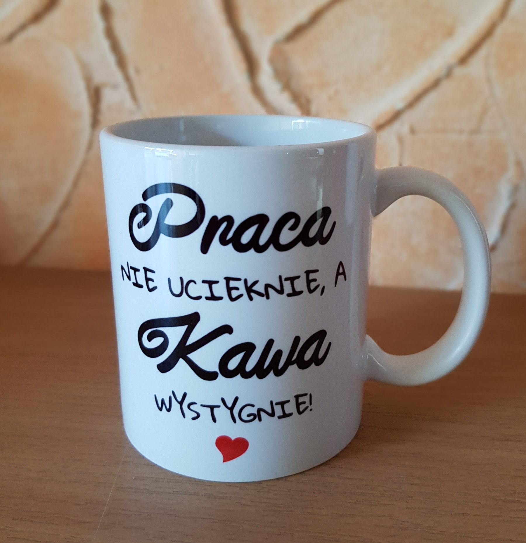 Biały kubek z czarnym napisem 'Praca nie ucieknie, a Kawa wystygnie!' i małym czerwonym sercem na tle ściany z beżowych kamieni.