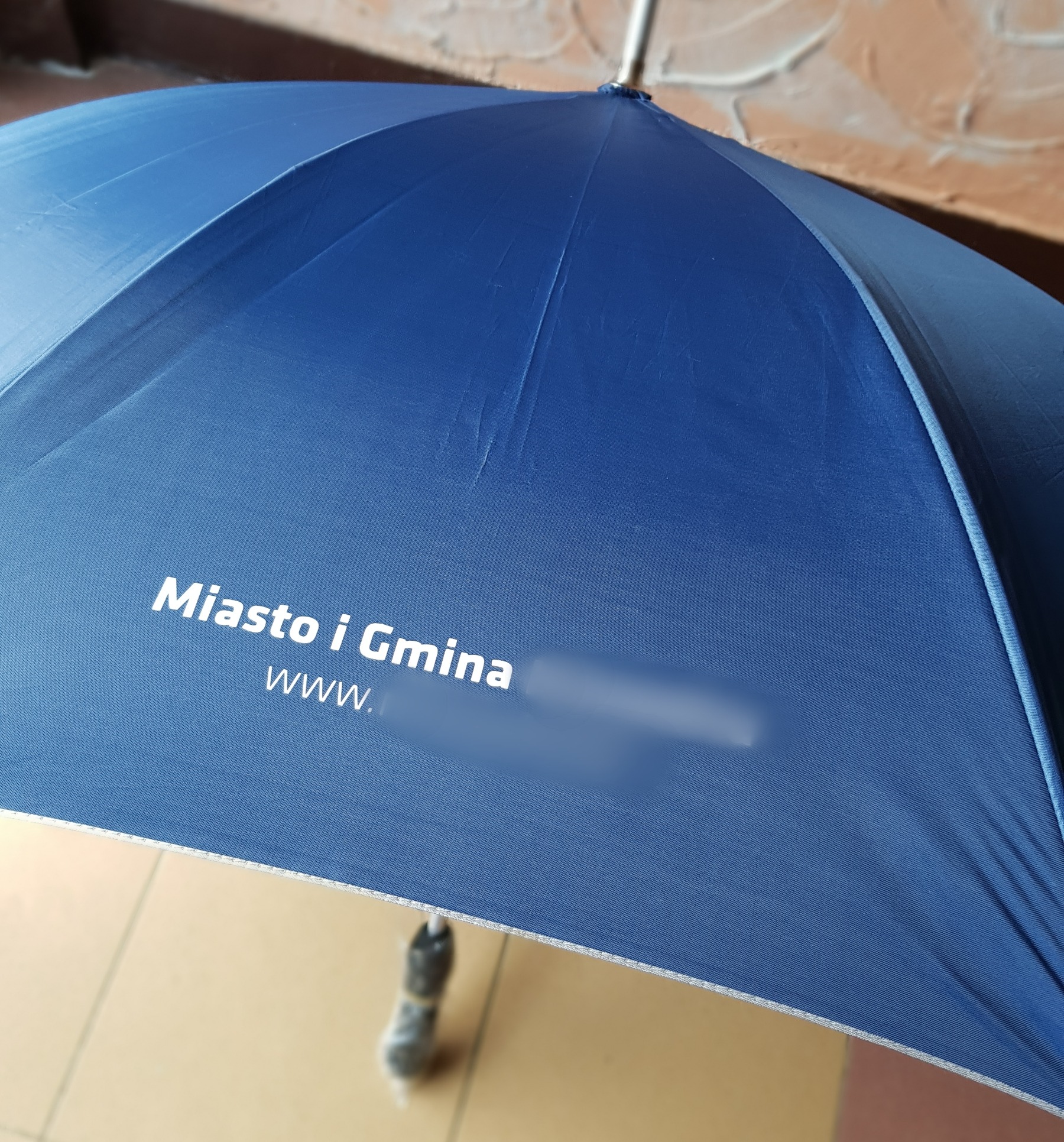 Niebieski parasol z białym nadrukiem 'Miasto i Gmina' oraz adresem strony internetowej z zamazaną nazwą domeny.