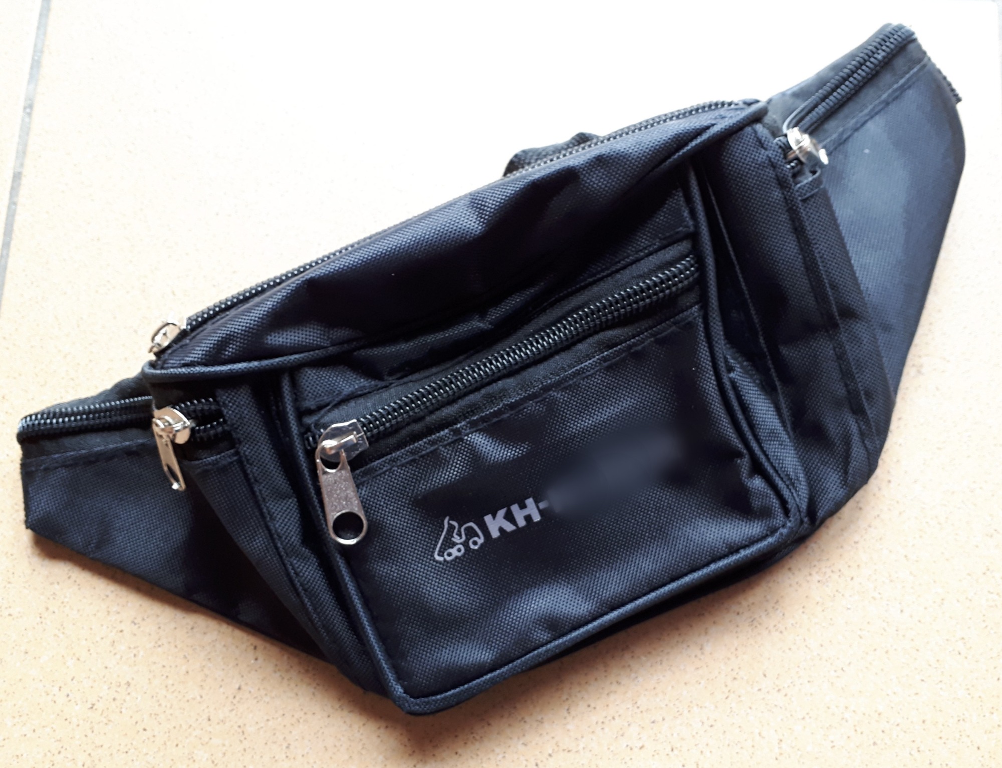 Czarna saszetka biodrowa z trzema zamkami błyskawicznymi i logo 'KH-' na tle beżowych płytek.