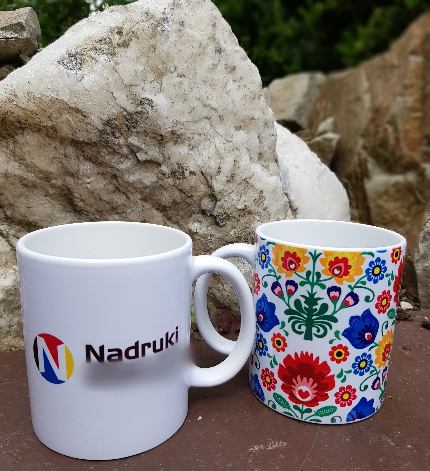 Dwa białe kubki z nadrukami, jeden z logo firmy 'Nadruki', drugi z kolorowym wzorem ludowym, na tle kamiennej ściany i zieleni.