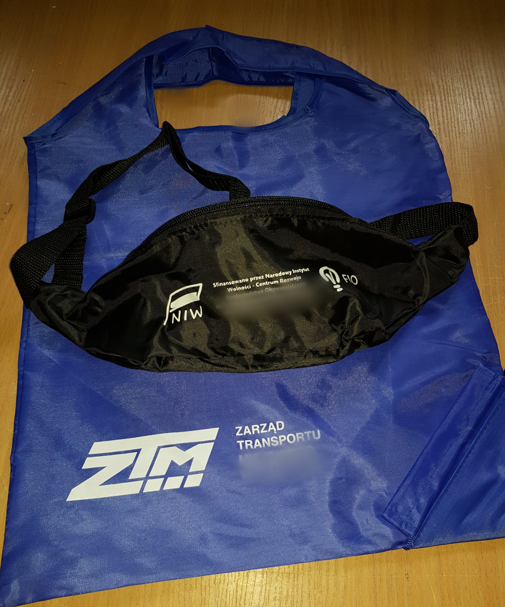 Czarna saszetka biodrowa z nadrukowanymi logotypami NIW i FIO leżąca na niebieskiej torbie z nadrukiem ZTM Zarząd Transportu.