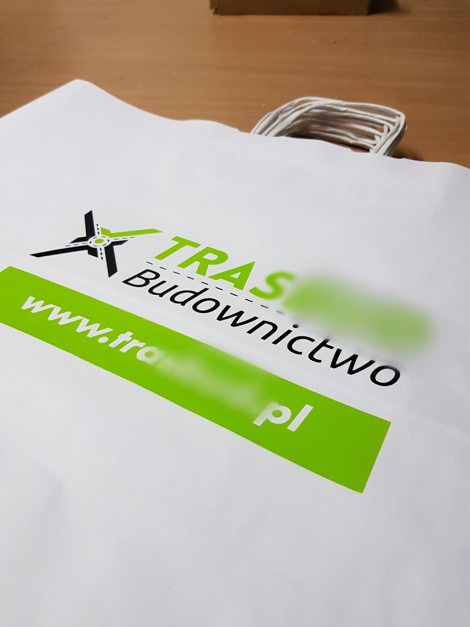 Biała torba papierowa z nadrukowanym logo firmy budowlanej w kolorze zielonym i czarnym, leżąca na drewnianym stole.