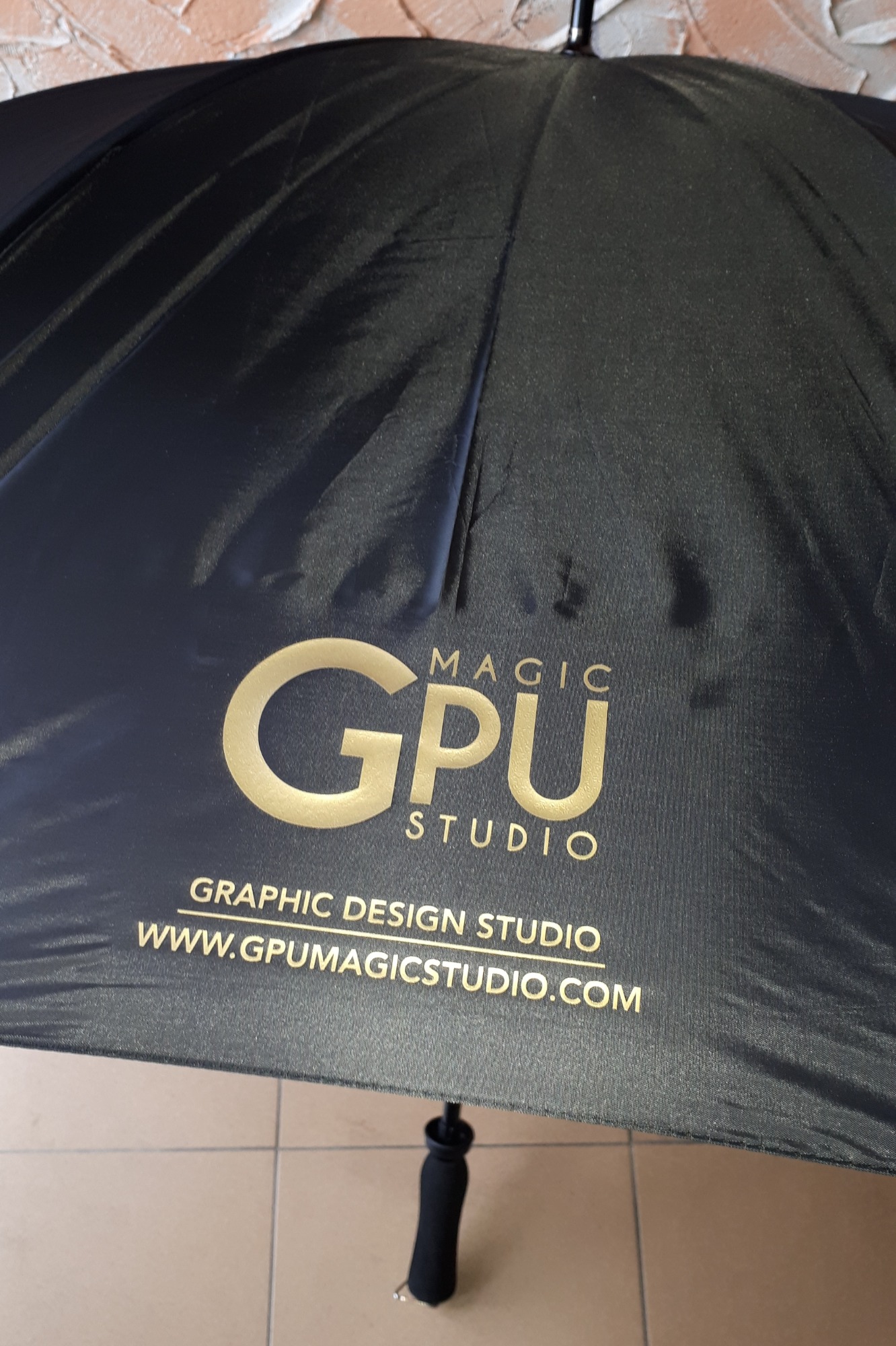Czarny parasol z nadrukowanym złotym logo 'GPU Magic Studio Graphic Design Studio www.gpumagicstudio.com', widoczne z bliska na tle beżowych płytek.