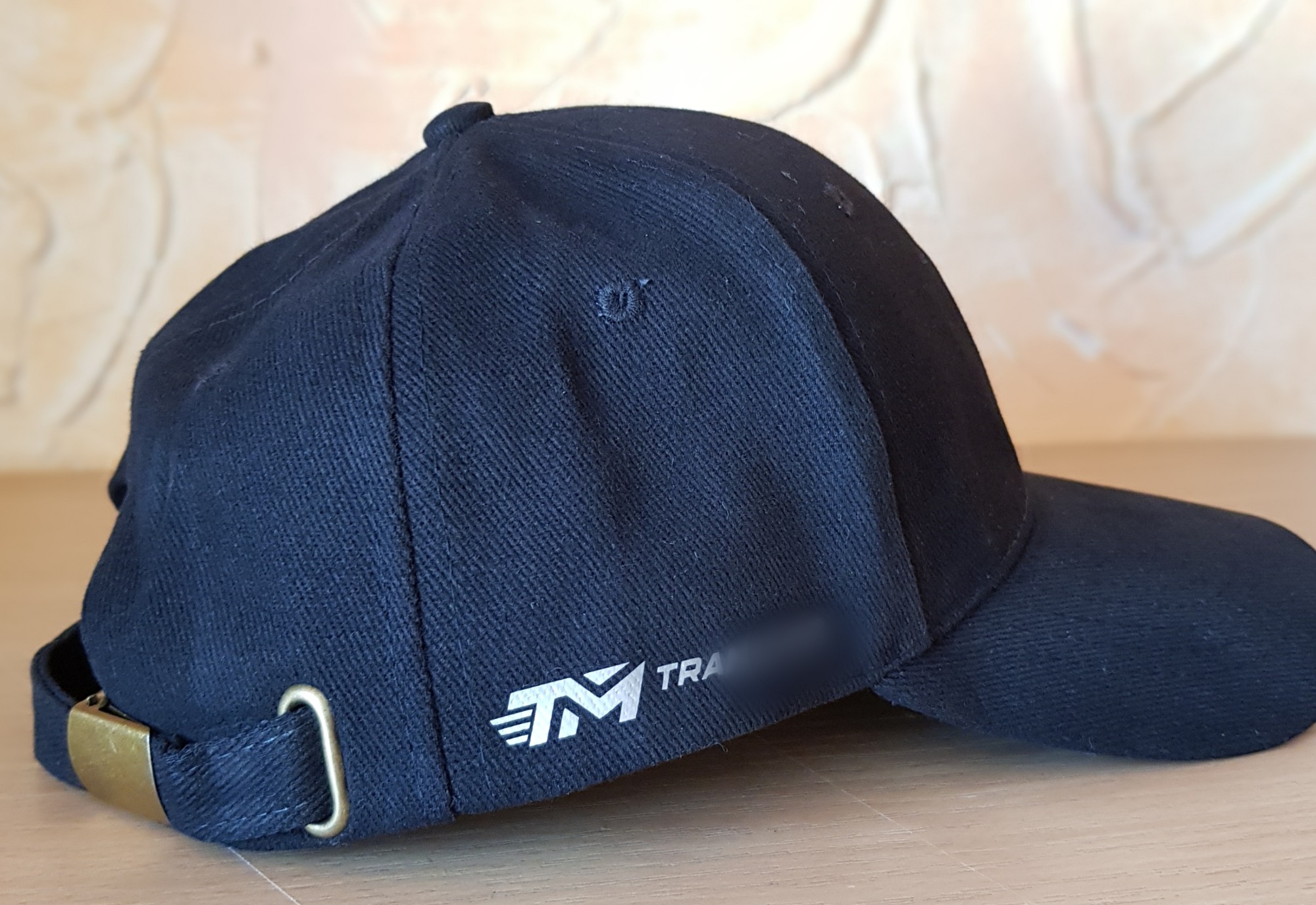 Czarna czapka z daszkiem z nadrukowanym białym logo firmy, leżąca na drewnianym blacie, widok z boku.