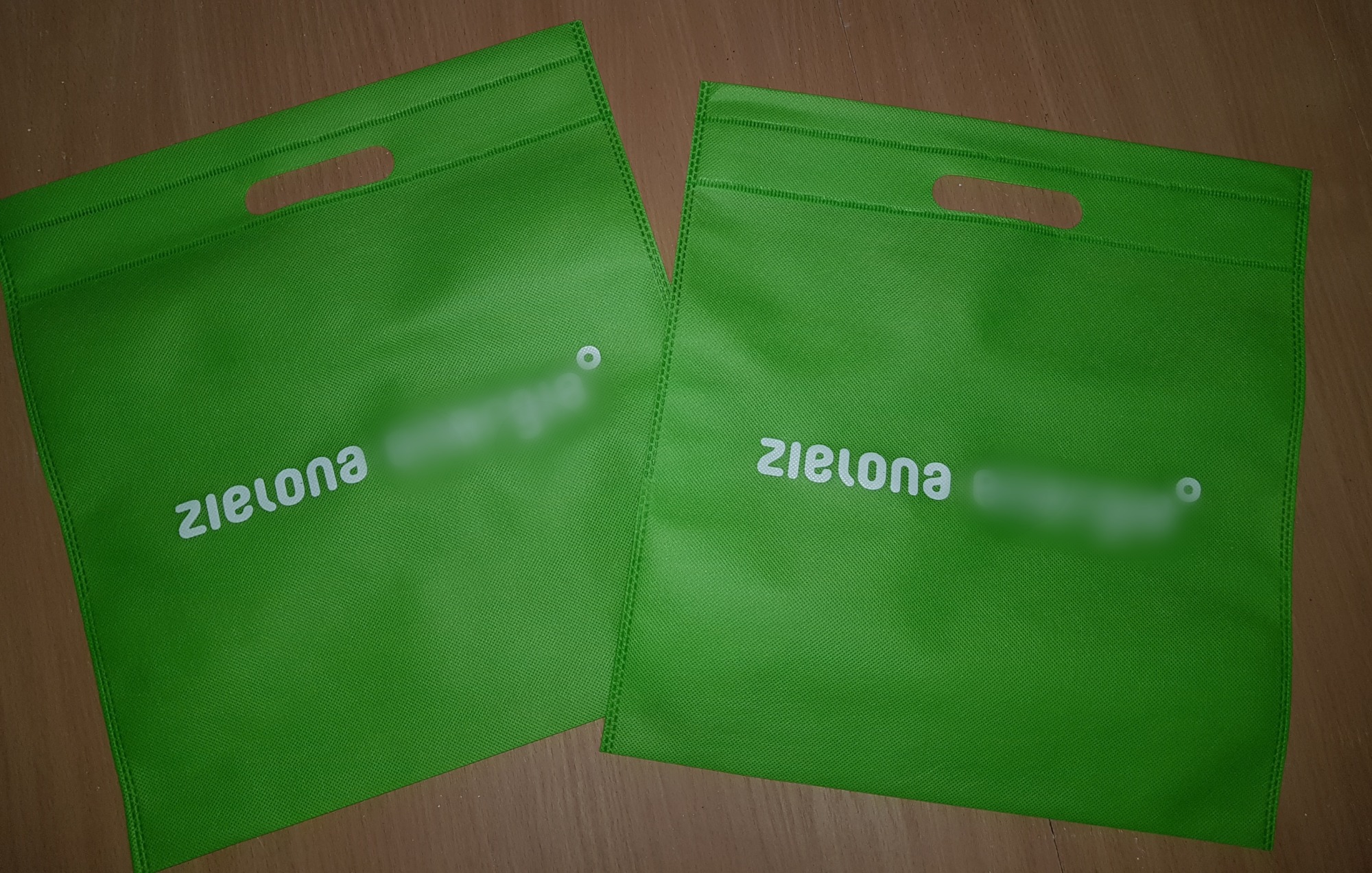 Dwie zielone, materiałowe torby z wyciętymi uchwytami i białym nadrukiem 'zielona' oraz zamazanym logo, leżące na drewnianym podłożu.