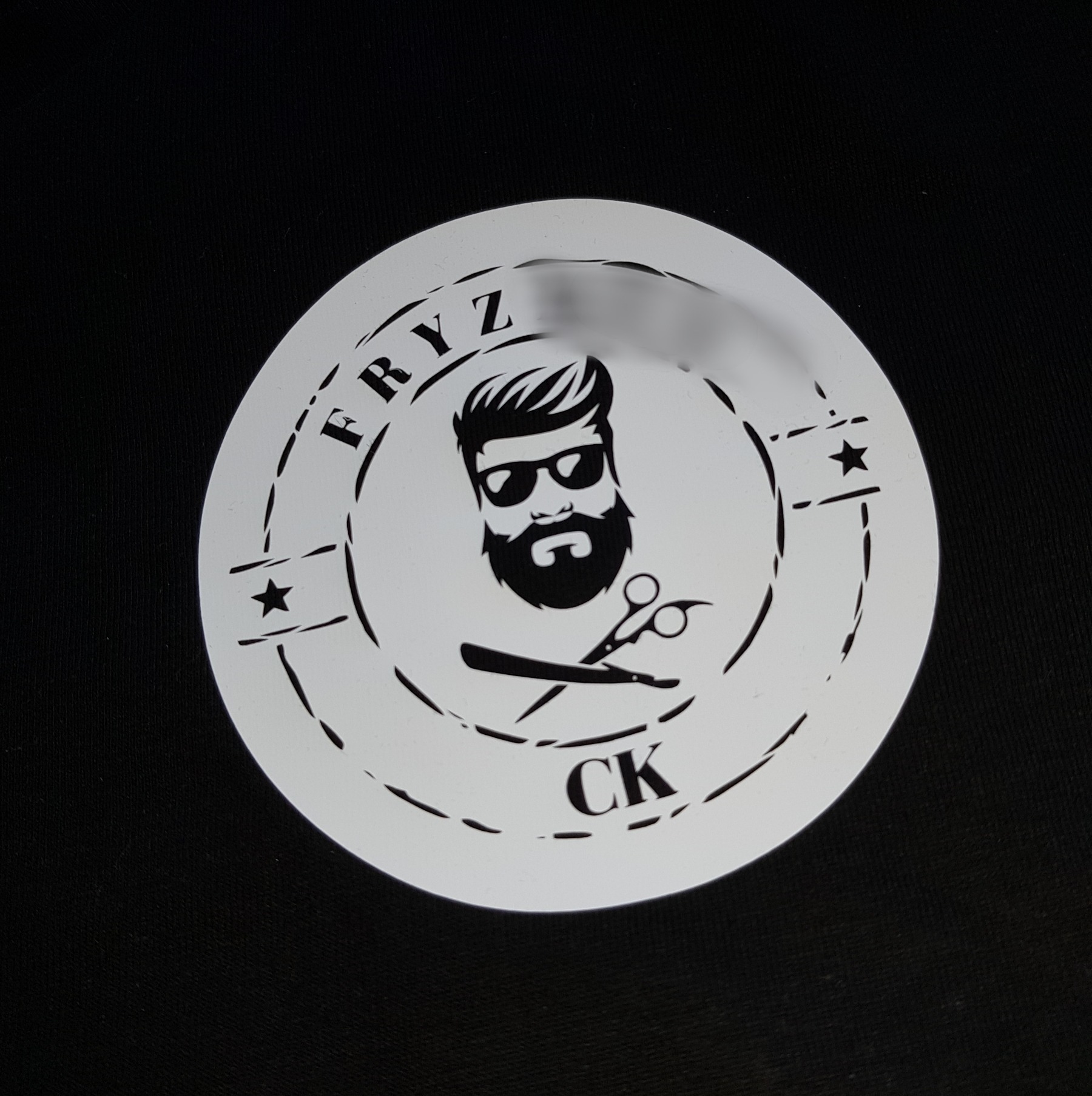 Szablon z wyciętym laserowo logo salonu fryzjerskiego przedstawiającego brodatego mężczyznę w okularach, nożyczki i brzytwę na czarnym tle.