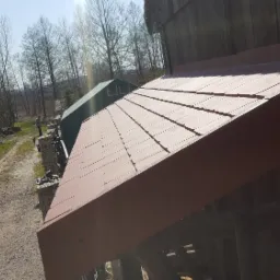 Czerwona dachówka na drewnianej konstrukcji z widocznymi drzewami w tle, ujęcie z niskiej perspektywy.