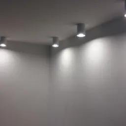 Minimalistyczne wnętrze z rzędem czterech okrągłych, montowanych natynkowo reflektorów LED, oświetlających gładką, szarą ścianę i sufit, tworząc subtelny efekt światłocienia.