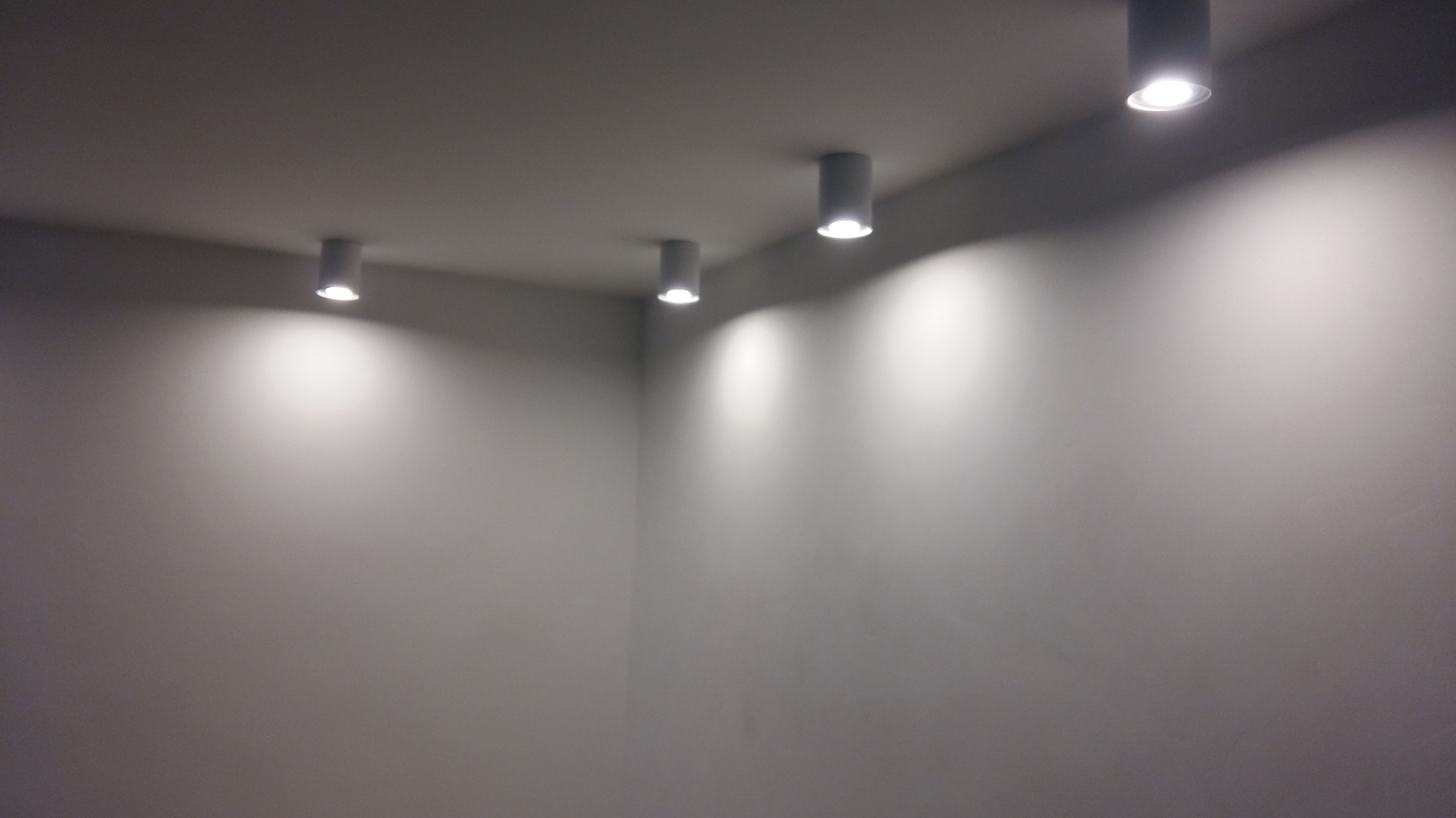 Minimalistyczne wnętrze z rzędem czterech okrągłych, montowanych natynkowo reflektorów LED, oświetlających gładką, szarą ścianę i sufit, tworząc subtelny efekt światłocienia.