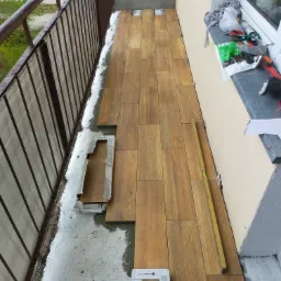 Montaż paneli podłogowych imitujących drewno na balkonie, widoczna poziomica, paczka z panelami i narzędzia na parapecie.