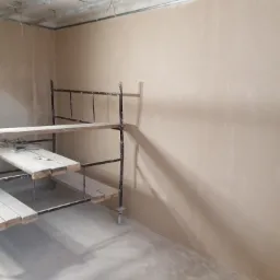 Świeżo otynkowane ściany w pomieszczeniu w trakcie remontu, widoczne rusztowanie z narzędziami na drewnianych deskach, metalowe profile sufitowe.