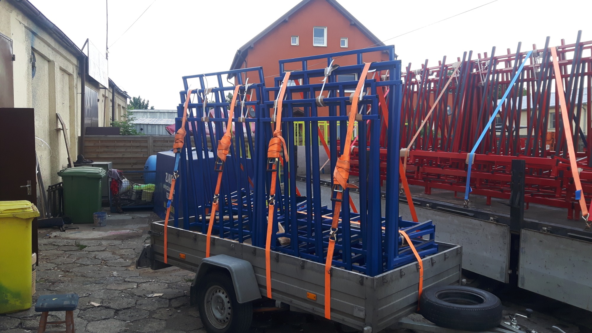 Przyczepa z niebieskimi konstrukcjami stalowymi zabezpieczonymi pomarańczowymi pasami transportowymi, za nią częściowo widoczna ciężarówka z czerwonymi konstrukcjami stalowymi, w tle budynki.