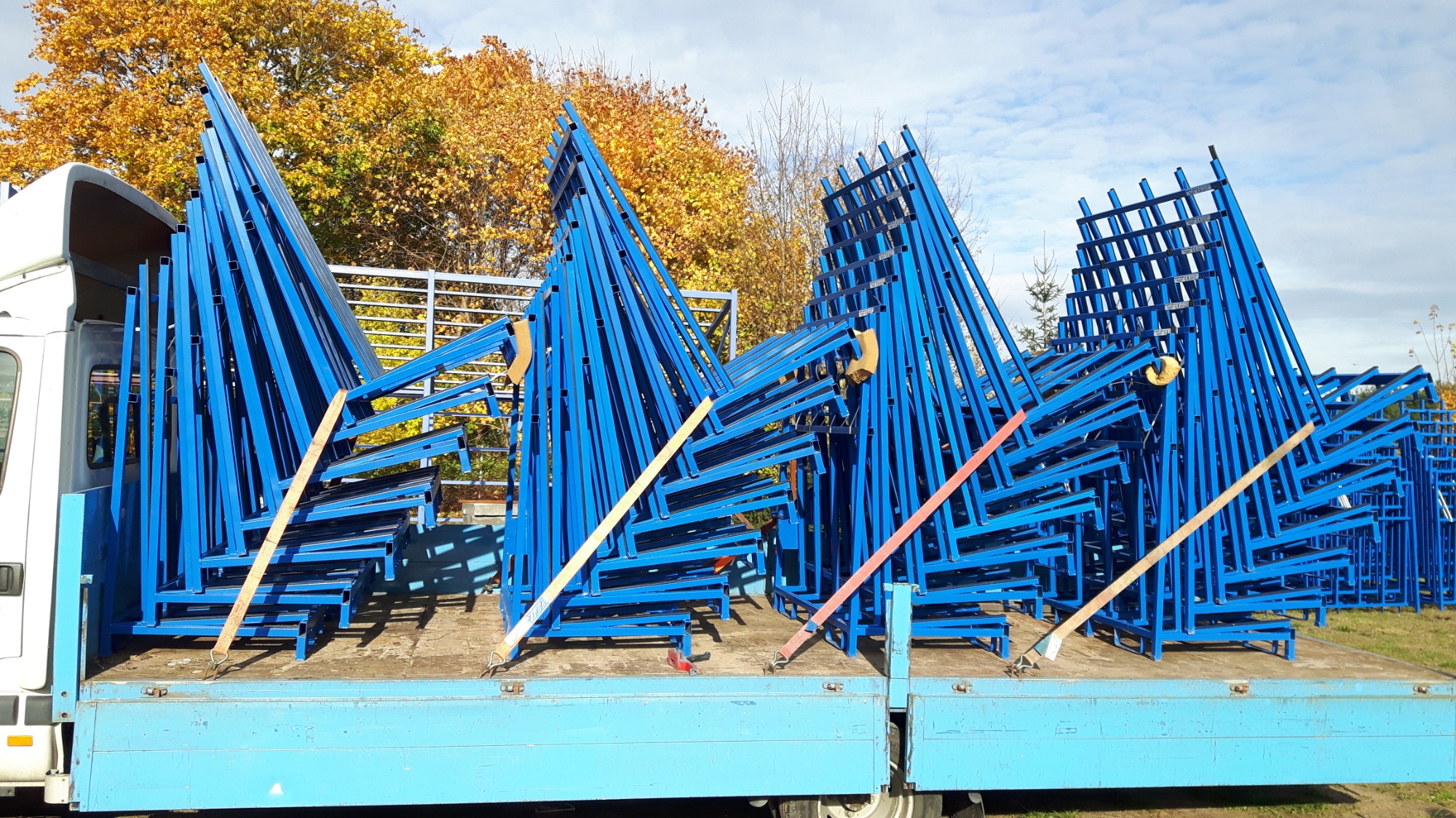 Niebieskie, metalowe konstrukcje halowe, zabezpieczone pasami transportowymi, przewożone na skrzyni samochodu dostawczego; w tle jesienne drzewa.