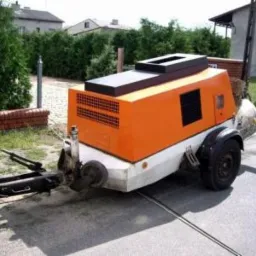 Pomarańczowo-biała maszyna do wylewania posadzek jastrychowych na przyczepie, z widocznym przewodem na szarej nawierzchni. Tło: budynki i zieleń.