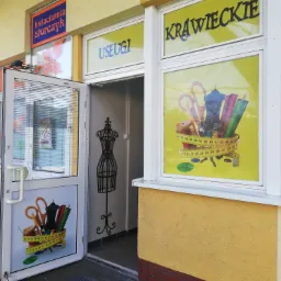 Wejście do zakładu krawieckiego z naklejkami na szybach przedstawiającymi nożyczki, manekin i miarę krawiecką. Widoczne napisy 'Krawieckie' i 'Usługi'.