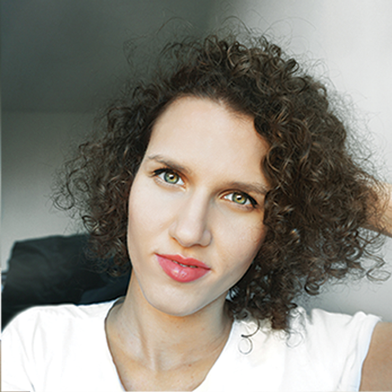 Portret kobiety o kręconych włosach, zielonych oczach i delikatnym makijażu, ubrana w białą koszulkę, z ręką uniesioną do włosów. Ujęcie z bliska, z lekkim uśmiechem.