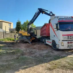 Żółta koparka kołowa Volvo załadowuje ziemię do czerwonej skrzyni białej ciężarówki Volvo na terenie budowy, w tle widoczny mężczyzna i budynek mieszkalny.