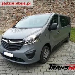 Szary bus Opel Vivaro z widocznym przodem i bokiem, zaparkowany na kostce brukowej przed kamiennym murem. Widoczne logo firmy Transmat.pl w prawym dolnym rogu.