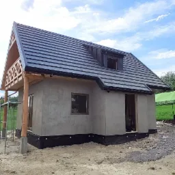 Surowy stan domu z drewnianą konstrukcją i szarą elewacją, z czarnym dachem i widocznym oknem dachowym, w trakcie budowy na piaszczystym terenie pod błękitnym niebem.