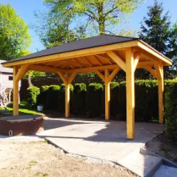 Drewniana altana ogrodowa z czarnym, skośnym dachem na betonowej kostce, otoczona zielenią, w słoneczny dzień.