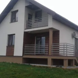 Dwupiętrowy dom z balkonem na piętrze i tarasem na parterze, oba zabezpieczone poziomymi, metalowymi barierkami; widoczne schody prowadzące na taras.