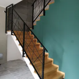 Schody drewniane z czarną, geometryczną balustradą metalową na tle turkusowej ściany i szarej podłogi.