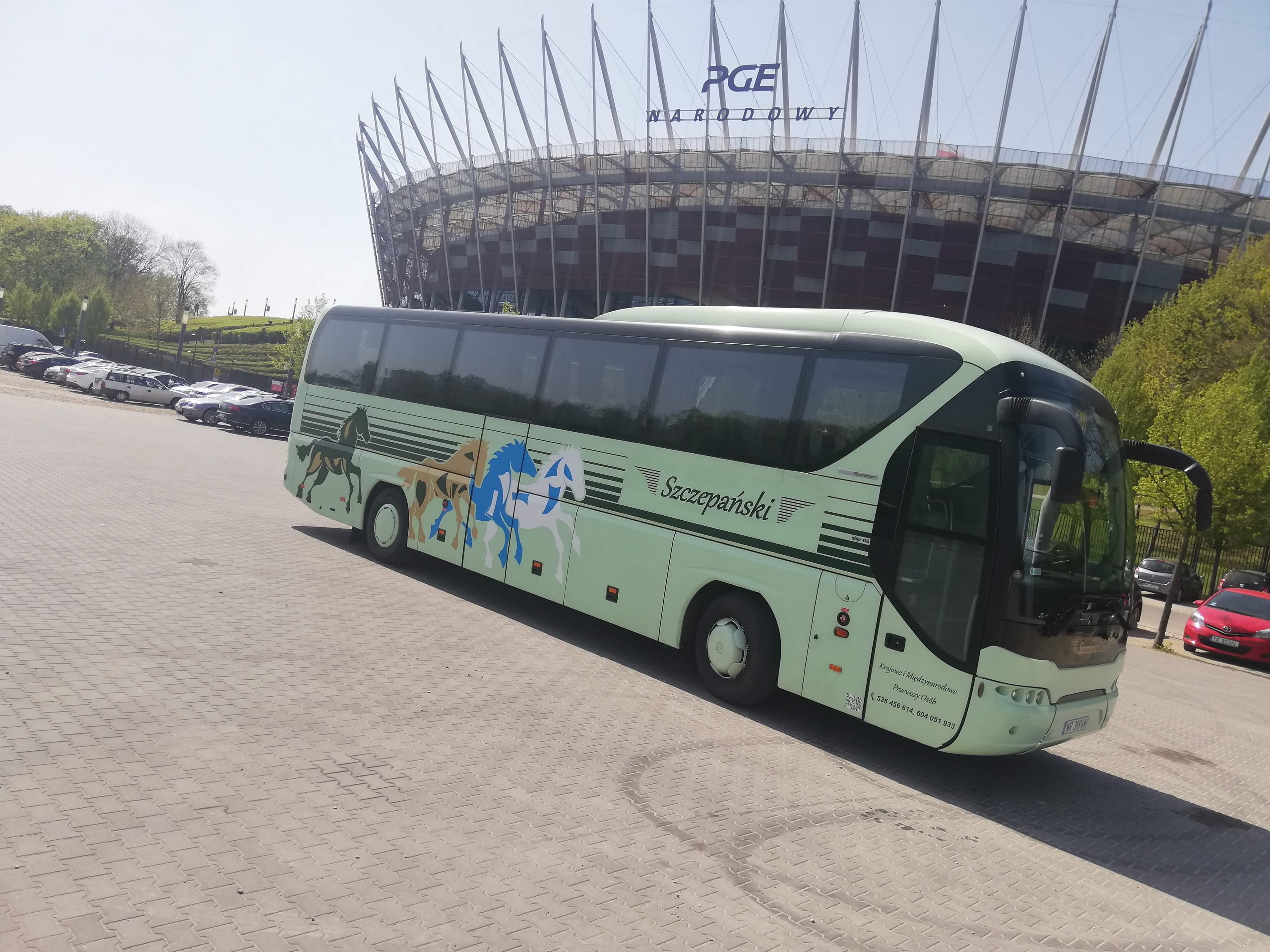 Autokar firmy Szczepański z grafiką koni na tle Stadionu Narodowego w Warszawie, zaparkowany na brukowanym placu.