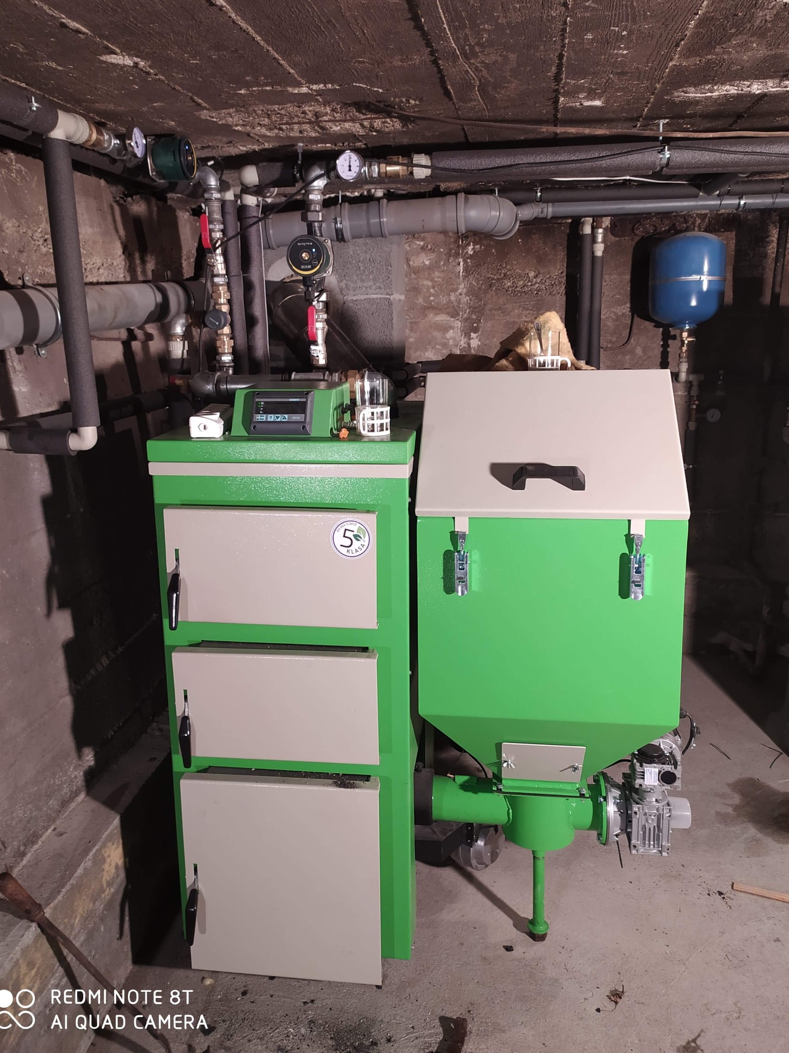 Nowoczesny piec na paliwo stałe, z podajnikiem, zainstalowany w kotłowni w Łapszach Wyżnych. Widoczne rury instalacji grzewczej.