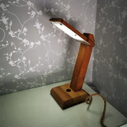 Drewniana lampka biurkowa z oświetleniem LED, stojąca na białym biurku na tle ściany z tapetą w kwiatowe wzory. Widoczny kabel w oplocie jutowym.