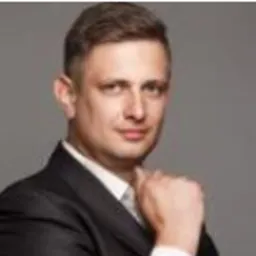 Portret mężczyzny w ciemnym garniturze na szarym tle, z ręką podpierającą podbródek. Uśmiech na twarzy.