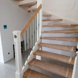 Nowe schody z jasnego drewna z zielonymi metalowymi balustradami, zabezpieczone folią ochronną podczas montażu, widoczne niedokończone prace elektryczne w tle.