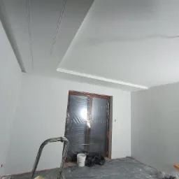 Świeżo pomalowane na biało wnętrze pokoju w Obszy z dekoracyjnymi listwami na suficie. Drzwi zabezpieczone folią, widoczna drabina malarska i akcesoria malarskie.