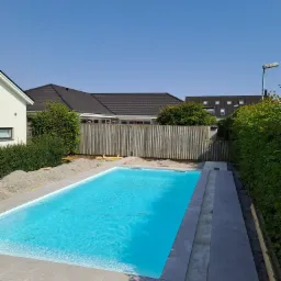 Niebieski basen w ogrodzie, obłożony szarymi płytami, widok na drewniany płot i domy w tle, prace wykończeniowe w toku.