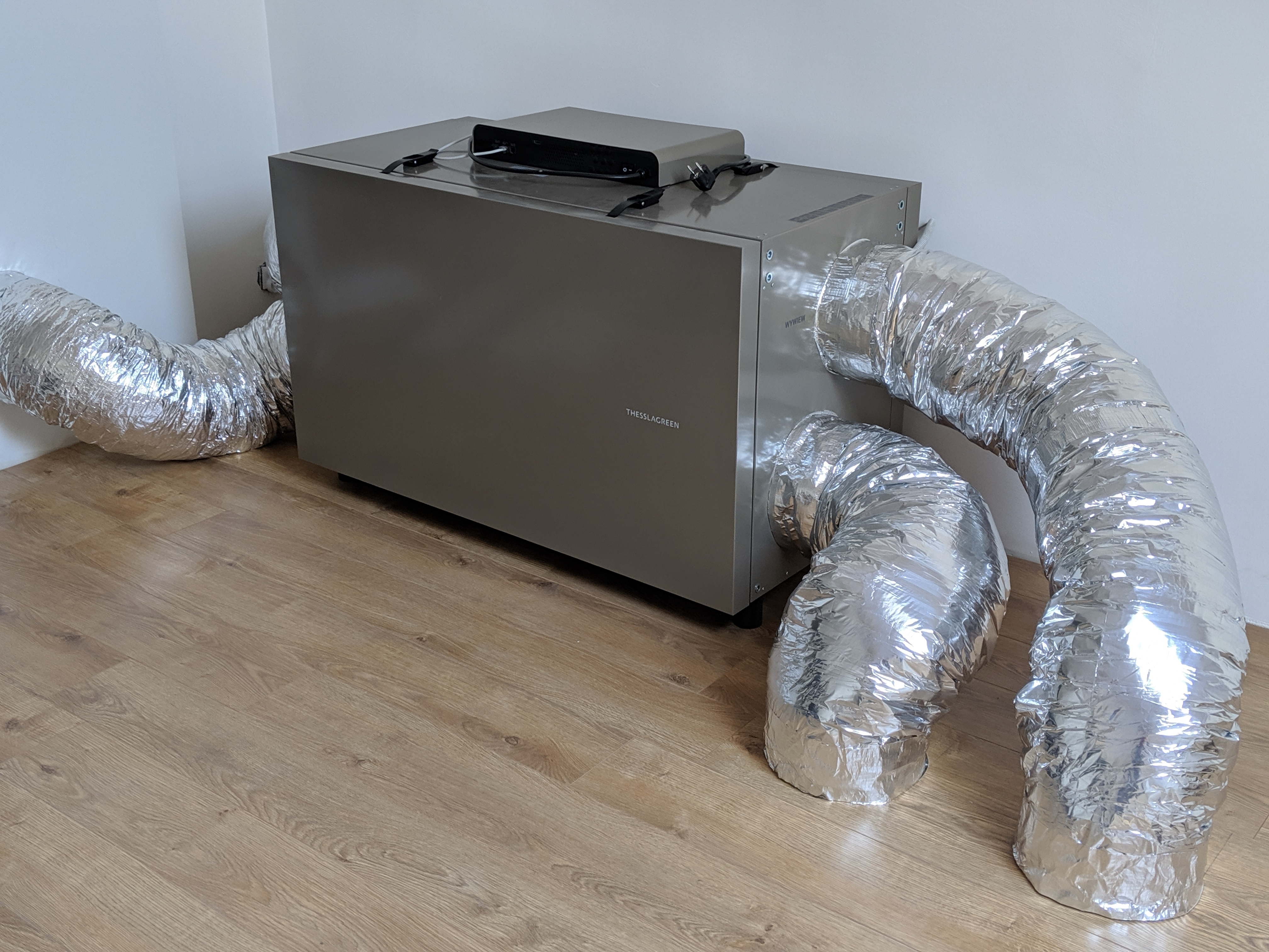 Szara jednostka klimatyzacyjna z podłączonymi elastycznymi przewodami wentylacyjnymi na drewnianej podłodze w rogu pokoju.