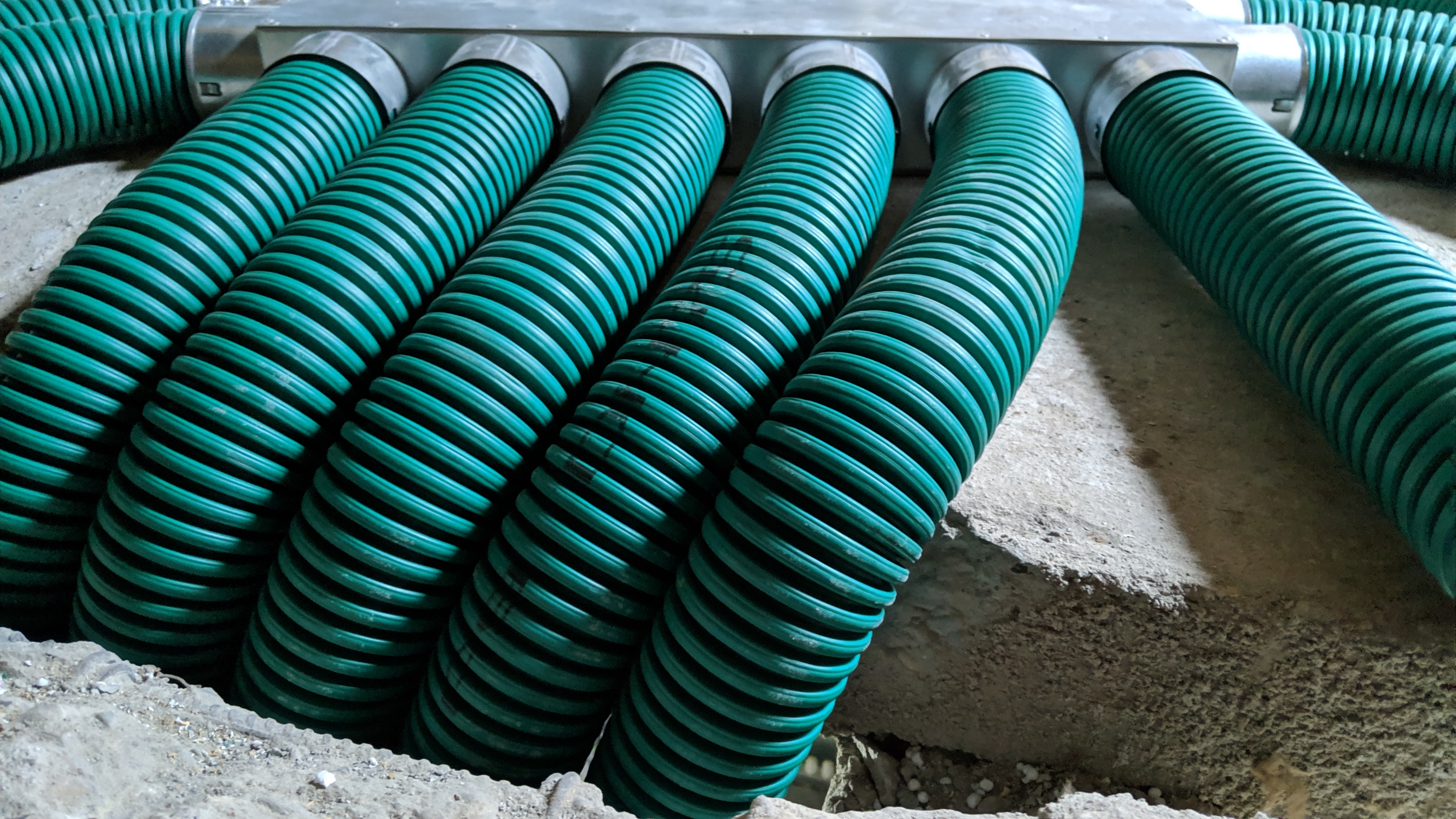 Szereg zielonych, karbowanych rur wentylacyjnych, zamocowanych do metalowej skrzynki rozdzielczej, wystających z betonowej podłogi w Muninie.