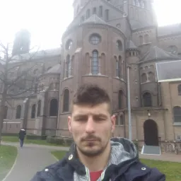 Mężczyzna na tle ceglanej architektury sakralnej w mglisty dzień. Widoczny fragment chodnika, trawnika i drzewa bez liści. Ujęcie z dołu, perspektywa żabia.