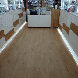 Kan-Bud Piotr Tomczak - Jasna podłoga z paneli w sklepie z oświetleniem LED przy regałach. Widok na wnętrze sklepu z perspektywy podłogi. Ciepłe światło podkreśla strukturę drewna.