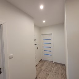 Kan-Bud Piotr Tomczak - Białe drzwi z poziomymi przeszkleniami w jasnym korytarzu z panelami podłogowymi imitującymi drewno. Minimalistyczny design, nowoczesne oświetlenie punktowe.