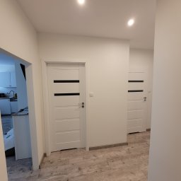 Kan-Bud Piotr Tomczak - Białe drzwi wewnętrzne z czarnymi akcentami w jasnym korytarzu z panelami podłogowymi imitującymi drewno. Widok na kuchnię w tle.