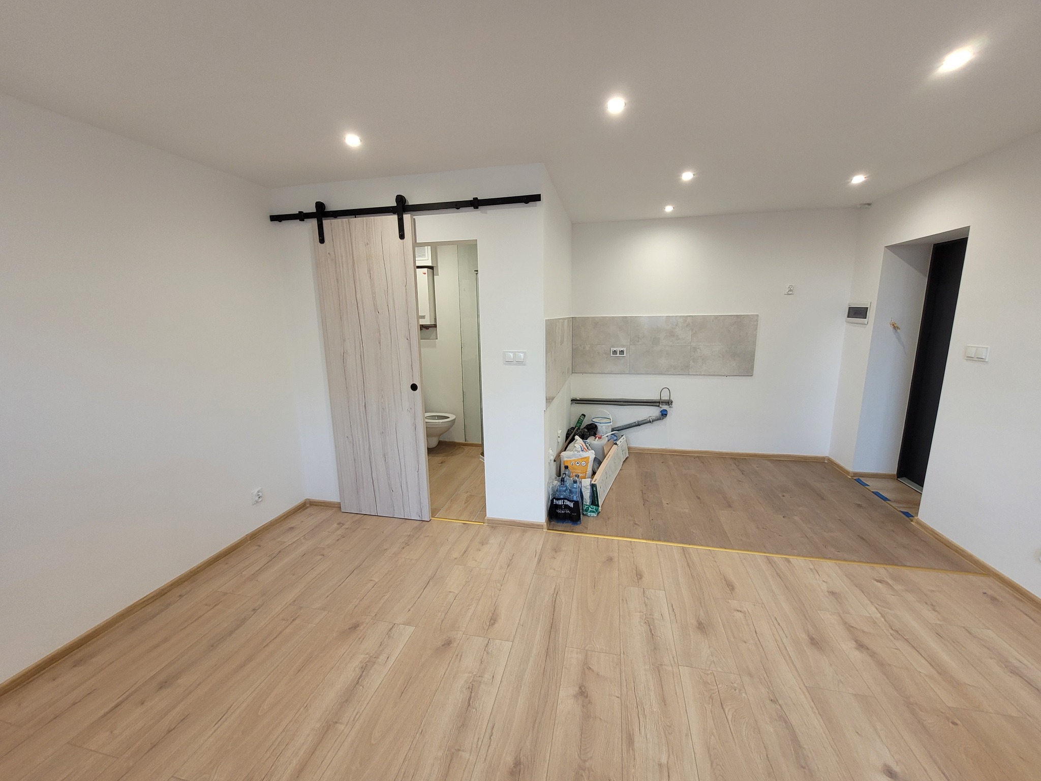 Jasne wnętrze po remoncie: panele podłogowe, przesuwne drzwi do łazienki, fragment kuchni z szarą glazurą. Widoczne materiały budowlane.