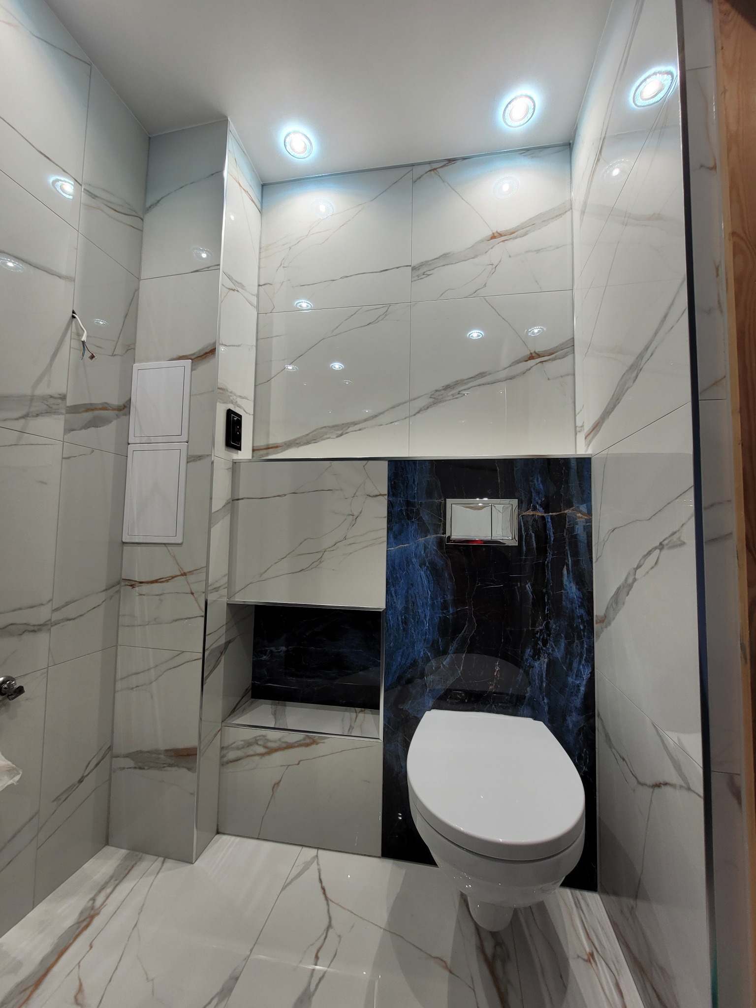Nowoczesna toaleta w wykończonym wnętrzu z białymi płytkami imitującymi marmur i akcentem z ciemnoniebieskiego kamienia za wiszącą muszlą klozetową.