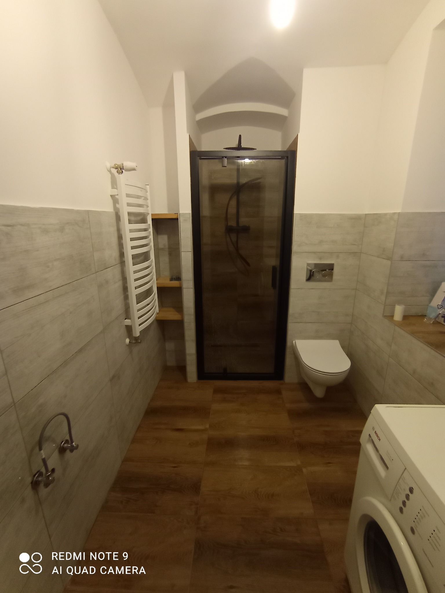 Nowoczesna łazienka po remoncie: kabina prysznicowa z czarną ramą, podwieszana toaleta, grzejnik drabinkowy i pralka Bosch. Płytki imitujące drewno na podłodze.