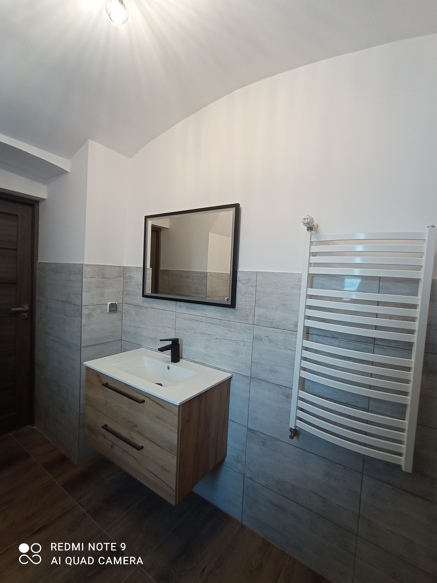 Nowoczesna łazienka po remoncie: drewniana szafka z umywalką, czarna bateria, lustro w czarnej ramie, biały grzejnik drabinkowy, szare płytki imitujące drewno.