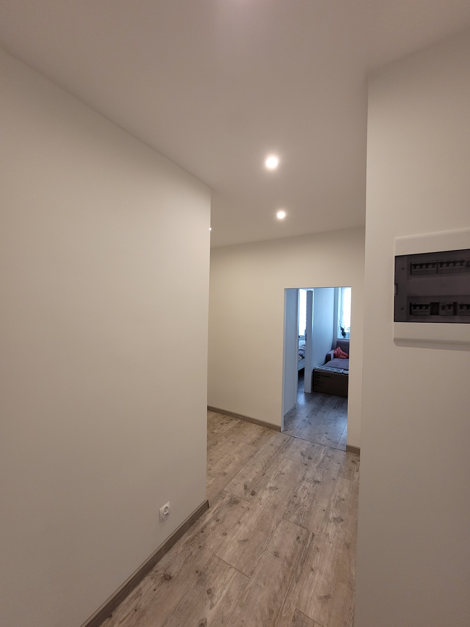 Jasny korytarz po remoncie z panelami podłogowymi imitującymi drewno, świeżo pomalowanymi ścianami i widoczną skrzynką elektryczną. W tle widok na pokój.