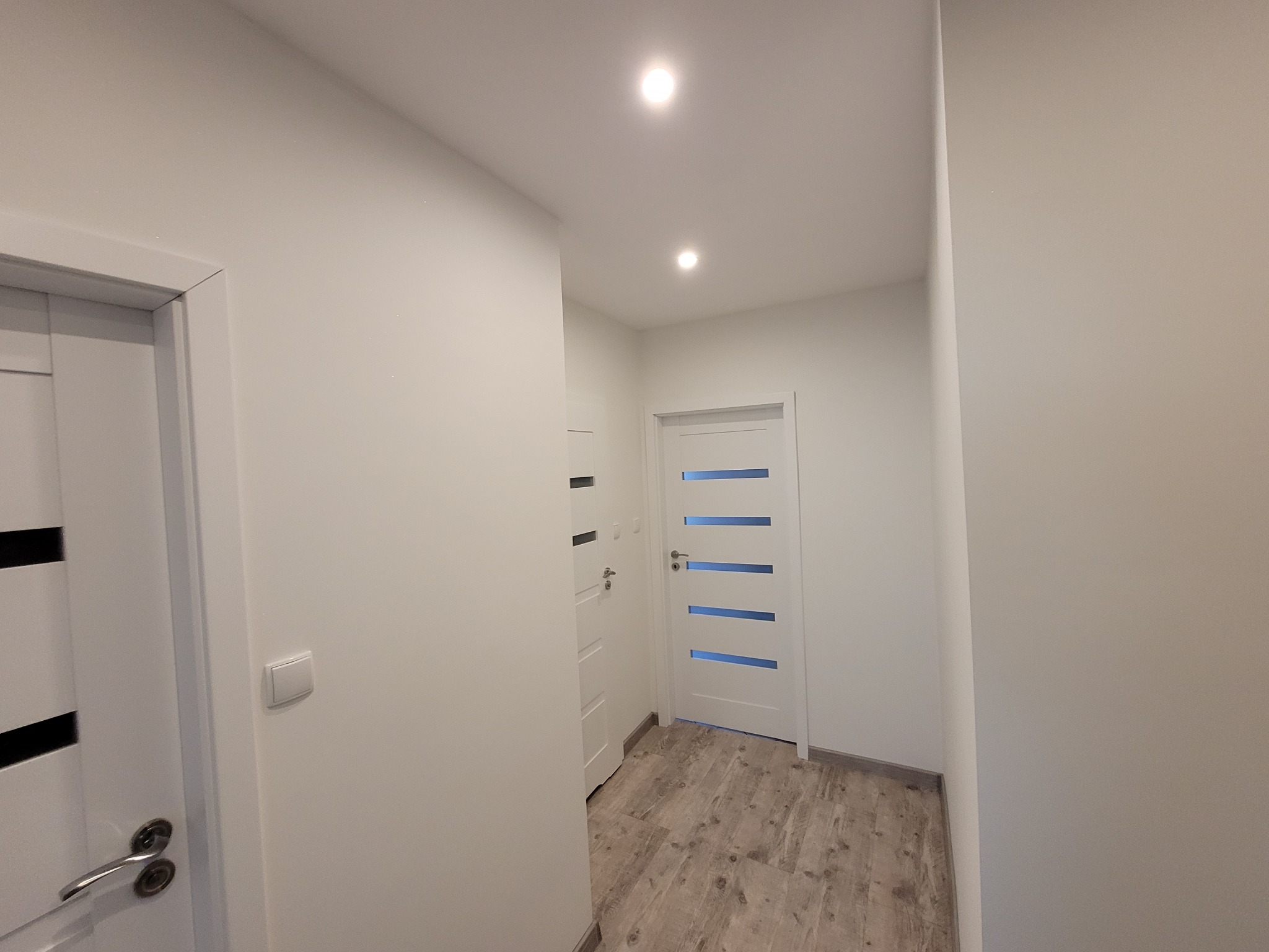 Białe drzwi z poziomymi przeszkleniami w jasnym korytarzu z panelami podłogowymi imitującymi drewno. Minimalistyczny design, nowoczesne oświetlenie punktowe.