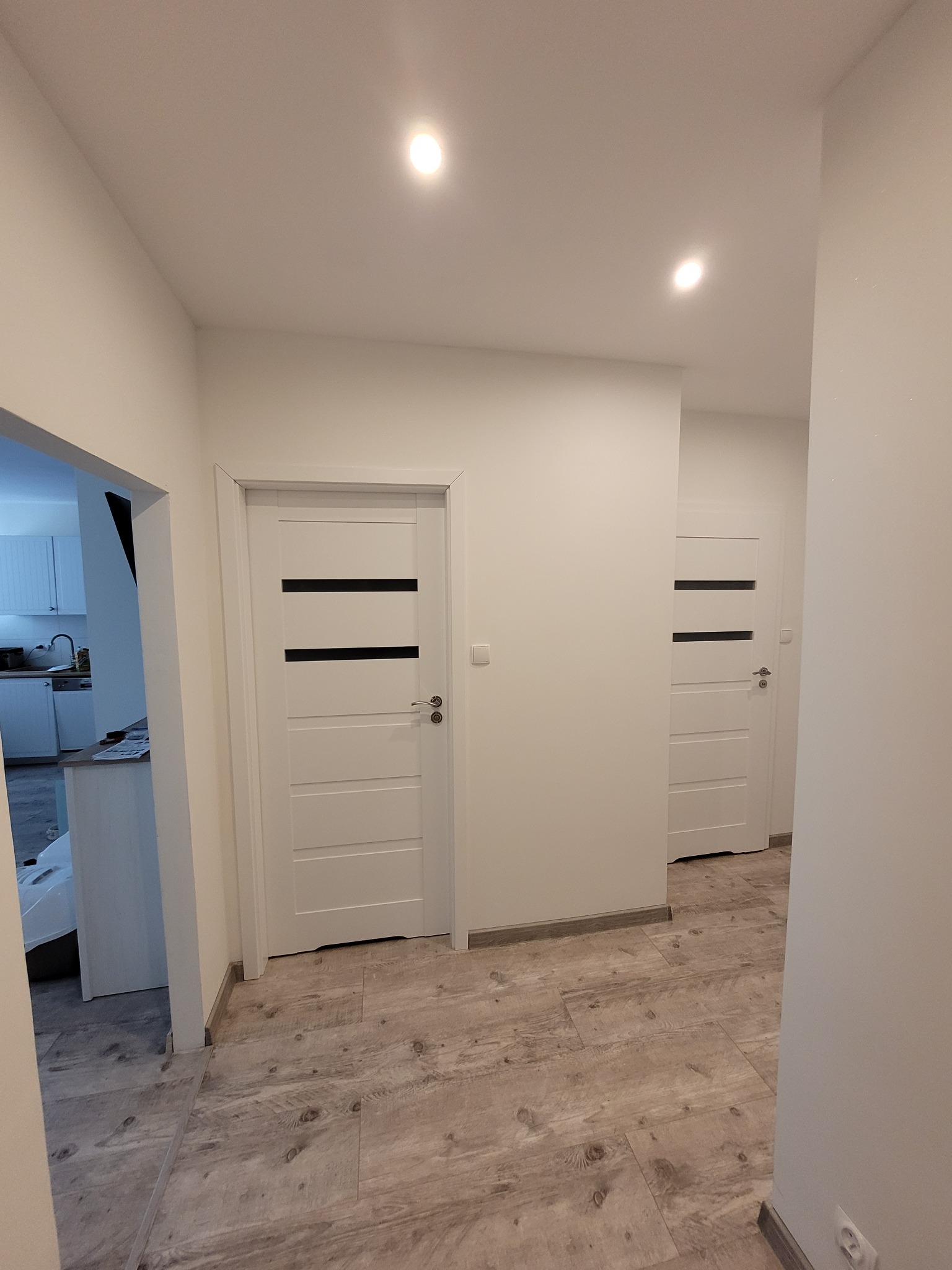 Białe drzwi wewnętrzne z czarnymi akcentami w jasnym korytarzu z panelami podłogowymi imitującymi drewno. Widok na kuchnię w tle.