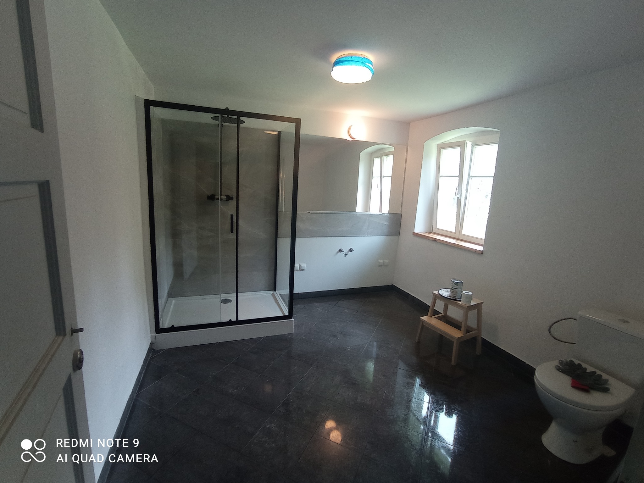 Wykończona łazienka z czarną kabiną prysznicową, ciemną podłogą i oknem z drewnianym parapetem. Na sedesie leżą rękawice robocze, a obok stoi stołek z farbą.