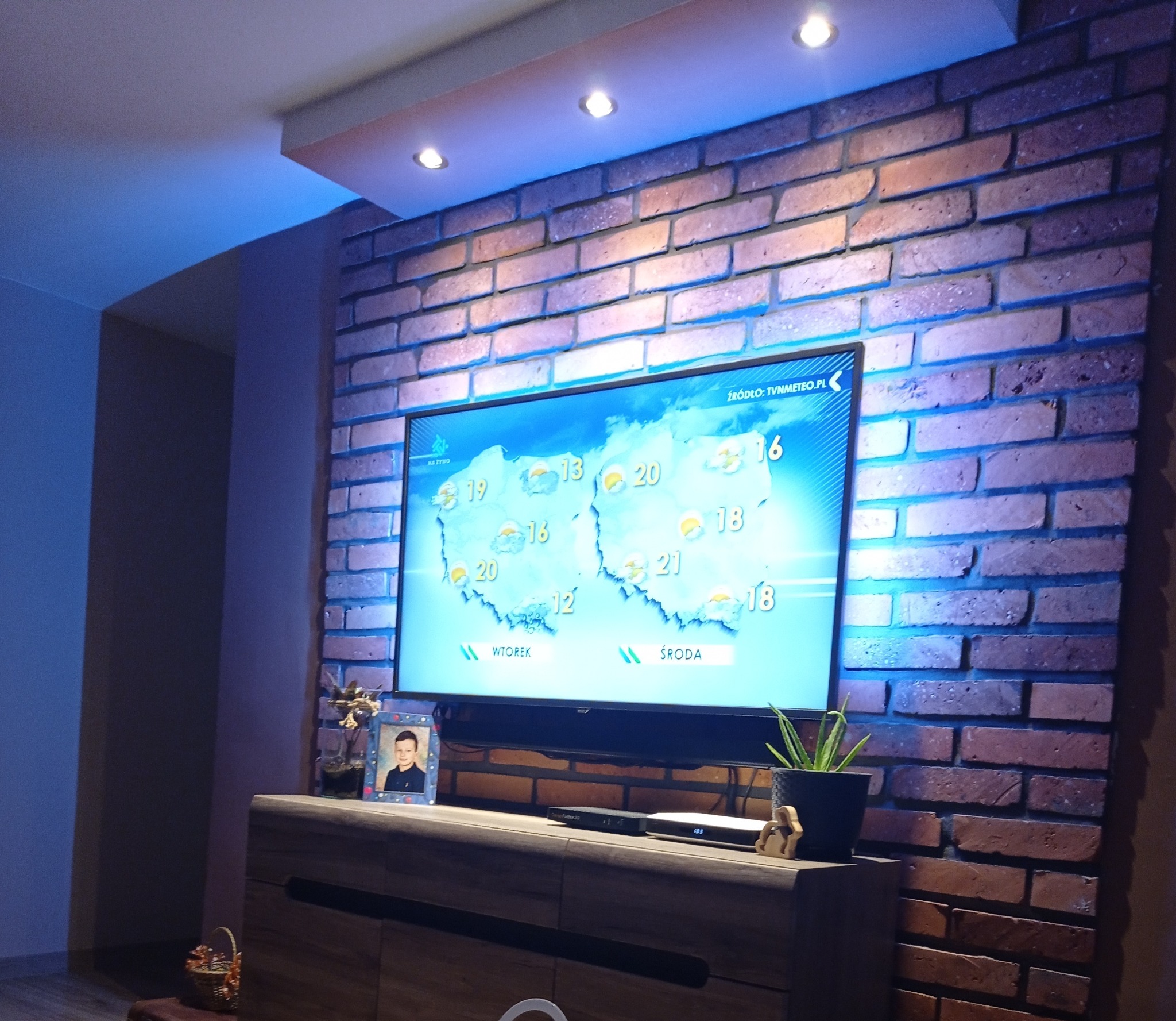 Ściana z cegły dekoracyjnej za telewizorem, podświetlona niebieskim światłem. Na szafce RTV ramka ze zdjęciem i roślina w doniczce. Nowoczesny design wnętrza.