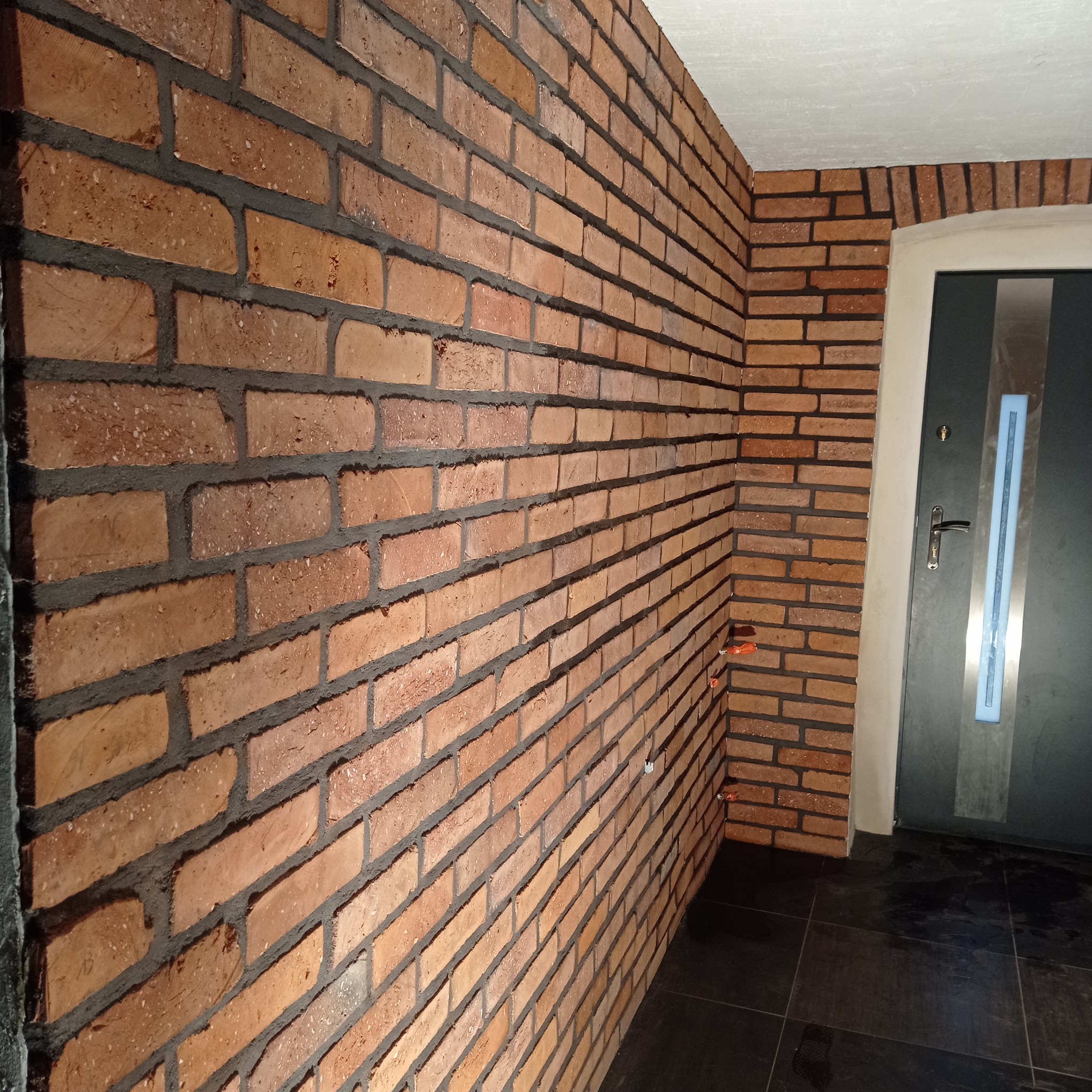 Ściana z cegły klinkierowej wewnątrz budynku we Wleniu. Widoczna szara fuga i fragment drzwi wejściowych. Wykończenie wnętrz w stylu industrialnym.